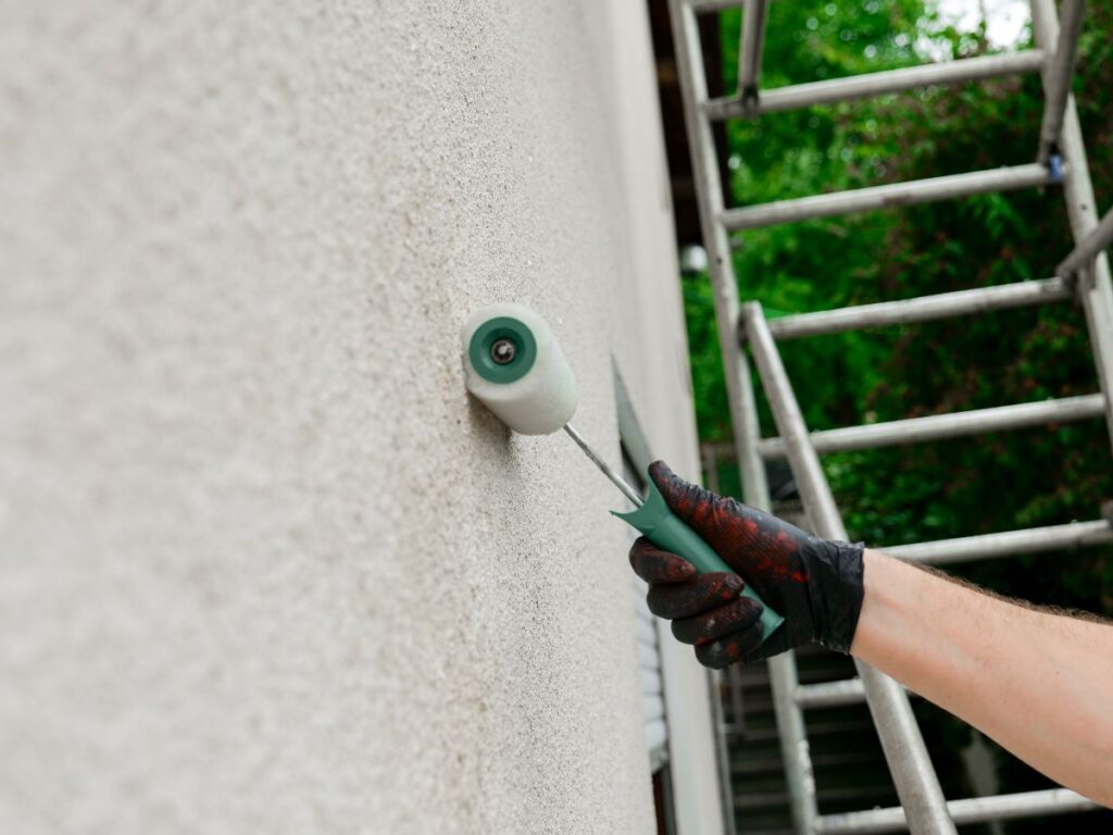 Hand using a paint roller on an exterior wall beside a ladder
